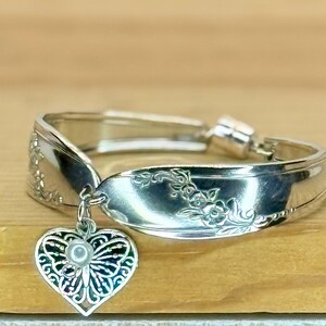 May include: A silver-toned spoon ring featuring an embossed floral pattern along the band. A heart-shaped charm with an intricate filigree butterfly design and a small white bead at the center hangs from the front of the metal band.