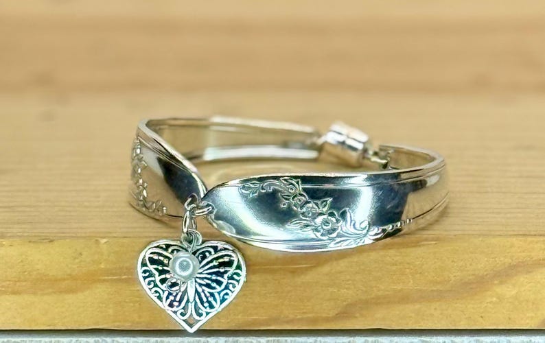 May include: A silver-toned spoon ring featuring an embossed floral pattern along the band. A heart-shaped charm with an intricate filigree butterfly design and a small white bead at the center hangs from the front of the metal band.