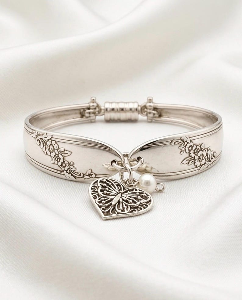 May include: A silver-toned metal cuff bracelet crafted from a repurposed spoon handle, featuring embossed floral patterns on the sides. The bracelet includes a dangling heart-shaped charm with an intricate butterfly cutout design and a small white pearl.