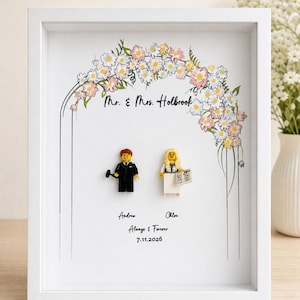 May include: A white framed shadow box featuring a printed floral arch with the text Mr. & Mrs. Holbrook. Two plastic minifigures are mounted inside. Below them, the text reads Andrew, Chloe, Always & Forever, and the date 7.11.2026.