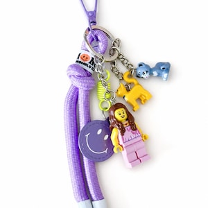 May include: A purple rope keychain featuring a purple plastic clip, a white tag with the text "I LOVE MONG LU 2", a purple circular smiley face charm, a yellow cat figure, a blue cat figure, and a plastic minifigure with brown hair.