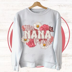 K&ouml;nnte beinhalten: Wei&szlig;er Pullover mit einem Blumendesign und dem Text "This Nana Prays".