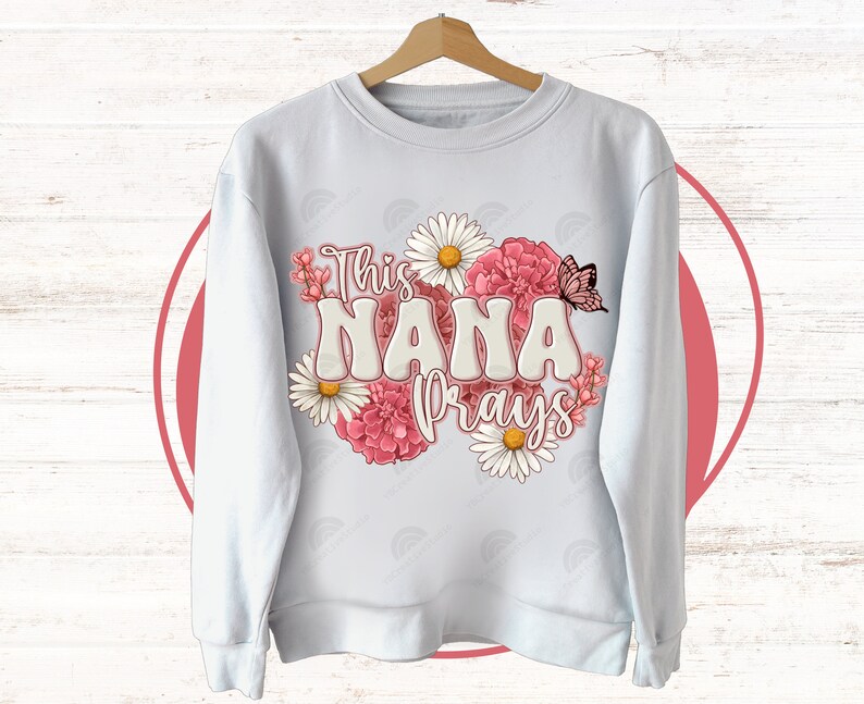 K&ouml;nnte beinhalten: Wei&szlig;er Pullover mit einem Blumendesign und dem Text "This Nana Prays".