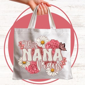 K&ouml;nnte beinhalten: Wei&szlig;e Canvas-Tragetasche mit rosa und wei&szlig;en Blumen und einem Schmetterling. Der Text "This Nana Prays" ist in Wei&szlig; auf die Tasche gedruckt.