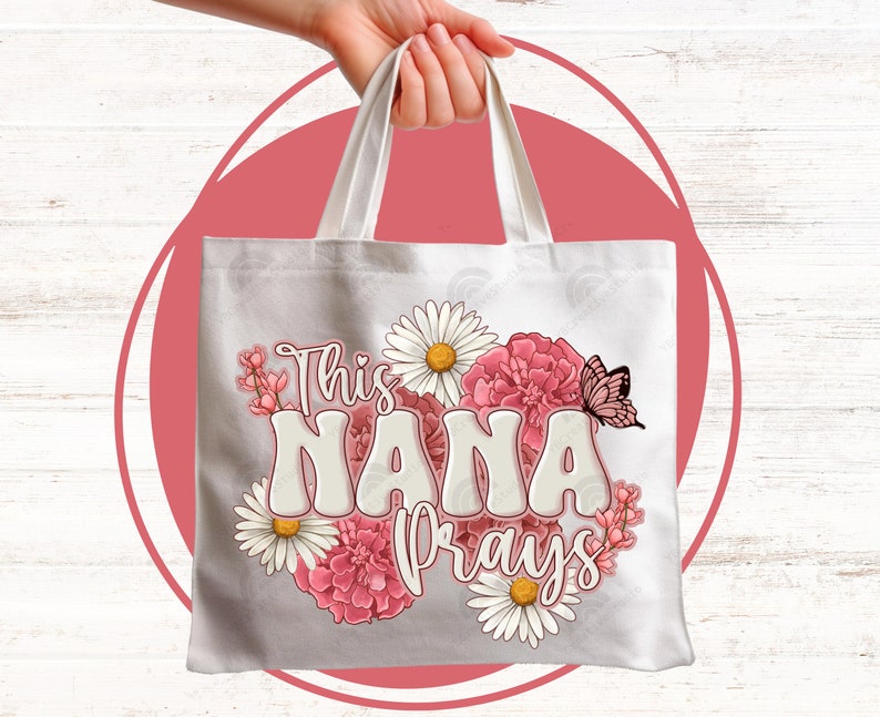 K&ouml;nnte beinhalten: Wei&szlig;e Canvas-Tragetasche mit rosa und wei&szlig;en Blumen und einem Schmetterling. Der Text "This Nana Prays" ist in Wei&szlig; auf die Tasche gedruckt.