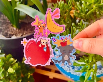May include: A set of four stickers featuring cartoon cats with different expressions. One sticker shows a cat with a banana, another shows a cat with an apple, and the other two show cats with different expressions. The stickers are printed on clear vinyl and have a glossy finish.