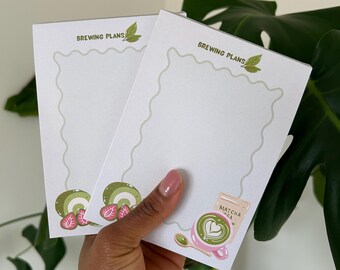 Może przedstawiać: Dwa białe notatniki z napisem "BREWING PLANS" i dekoracyjnymi zielonymi liśćmi. Notatniki mają falisty brzeg i ilustracje herbaty matcha i plasterków kiwi. Notatniki są trzymane przez osobę.