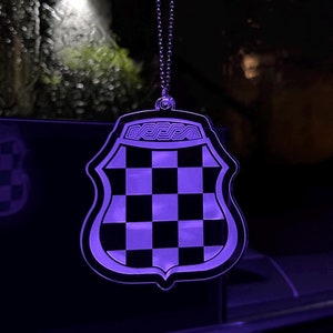 May include: A purple-lit, shield-shaped car charm with a black and white checkered pattern. The charm is suspended by a silver chain. The top of the shield has the word "DEDCA" in a stylized font. The background is dark.