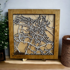 Puede incluir: Un mapa enmarcado en madera, con un detallado diseño de una ciudad cortado con láser en blanco sobre un fondo marrón claro. La palabra "Livno" está escrita en cursiva. El marco es de madera natural.