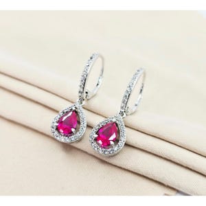 May include: A pair of silver-toned drop earrings. Each earring features a teardrop-shaped, vibrant red gemstone surrounded by a halo of small, clear crystals. The earring's hoop is also encrusted with clear crystals, adding sparkle.
