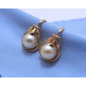 May include: A pair of gold-colored earrings featuring large, round, white pearls. The earrings have a teardrop-shaped design with delicate leaf accents. The earrings are set against a light blue background.