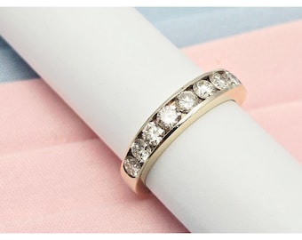 May include: A gold band ring with multiple round, clear gemstones set in a channel. The ring is displayed on a white cylindrical object against a pink and blue background. The gemstones sparkle.