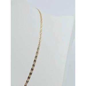 May include: A gold-colored necklace with a delicate, repeating pattern of rectangular links. The necklace is displayed on a white mannequin, showcasing its design and length. The links have a textured, starburst-like design.