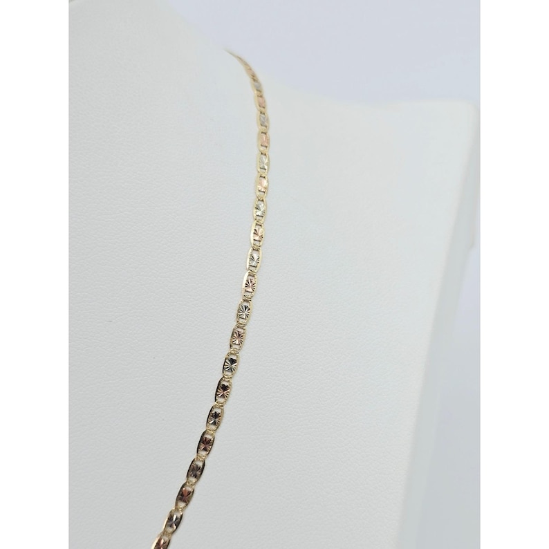 May include: A gold-colored necklace with a delicate, repeating pattern of rectangular links. The necklace is displayed on a white mannequin, showcasing its design and length. The links have a textured, starburst-like design.