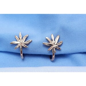 May include: A pair of gold-colored clip-on earrings, each shaped like a stylized cannabis leaf. The earrings are displayed on a light blue fabric background. The earrings have a shiny, metallic finish.