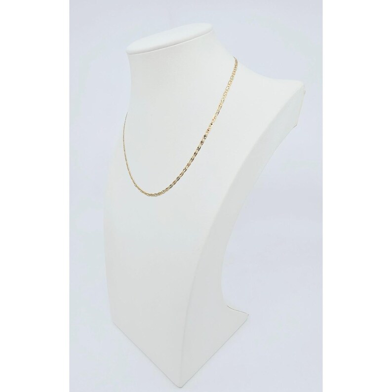 May include: A delicate gold-colored chain necklace displayed on a white mannequin bust. The necklace has a repeating pattern of small, rectangular links. The bust is set against a plain white background, highlighting the jewelry.