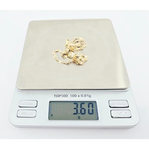 May include: A gold-colored chain necklace rests on a digital scale. The scale displays a weight of 3.60 grams. The scale is silver with the text "TOP500 500 x 0.01g".