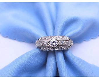 May include: A silver ring with a central diamond and surrounding smaller stones. The ring is set against a light blue fabric background, creating a contrast that highlights the ring's intricate design and sparkle. The ring's design features geometric shapes.