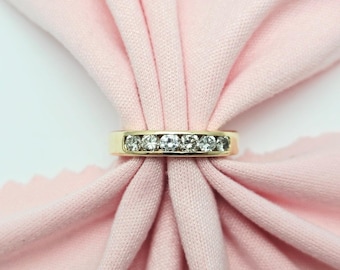 May include: A gold band ring with a row of clear, sparkling gemstones. The ring is set against a soft pink fabric background, creating a contrast between the gold and the gemstones.