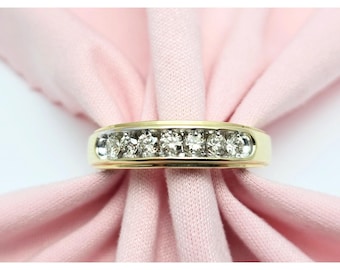 May include: A gold band ring with seven clear, round-cut diamonds set in a row. The ring is displayed on a light pink fabric, creating a contrast with the bright gemstones and the gold setting. The ring is a classic design.