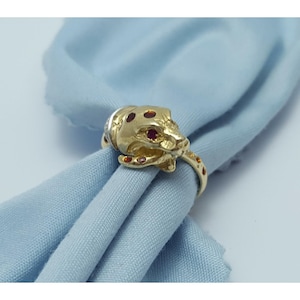 May include: A gold-colored ring featuring a jaguar head design with red gemstone accents. The ring is set against a light blue fabric background, showcasing its intricate details and craftsmanship. The ring's band is also adorned with small red gemstones.