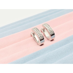 May include: A pair of silver hoop earrings with a row of small, clear stones set along the front. The earrings are displayed on a pink and blue striped surface. The earrings have a polished, reflective finish.