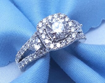 May include: A diamond engagement ring with a heart-shaped halo setting. The ring features a large, round center stone surrounded by smaller diamonds. The band is also encrusted with diamonds, creating a sparkling effect. The ring is set on a light blue fabric.