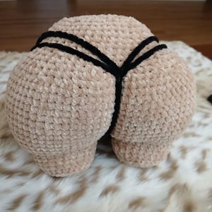 May include: A plush, beige crocheted buttock-shaped pillow with black string detailing. The pillow is made of a soft, textured material and is displayed on a fluffy, patterned surface.
