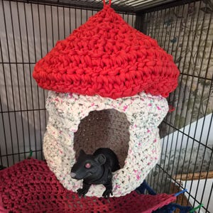 May include: A crocheted hanging pet hideout shaped like a mushroom with a bright red cap and a white base featuring a small floral pattern. A black toy rat is positioned inside the opening of the structure, which is suspended within a wire cage.