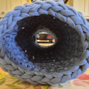 May include: A cylindrical tube crafted from light blue fabric yarn, featuring a thick braided edge at the opening and a dense, textured crochet stitch pattern throughout the interior walls, creating a hollow, tunnel-like structure.