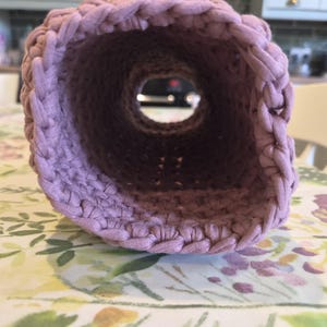 May include: A cylindrical, mauve-colored crochet tube crafted from thick fabric yarn. The item features a consistent, textured stitch pattern throughout its length, forming a hollow, open-ended structure with a circular opening at the front.