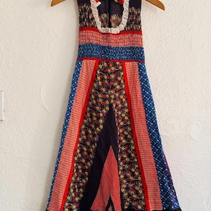 May include: A vintage sleeveless dress with a patchwork design. The dress features a mix of floral, striped, and dotted patterns in red, blue, and black. It has a ruffled hem and a white lace collar. The dress is hanging on a wooden hanger.