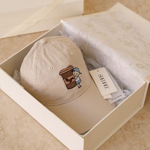 Puede incluir: Gorra de béisbol beige con un diseño bordado de una taza de café y un personaje de dibujos animados. Presentada en una caja de regalo blanca con papel de seda. Una etiqueta de marca está adjunta.
