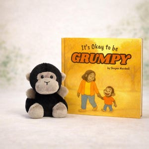 Może przedstawiać: Pluszowa czarno-szara zabawka w kształcie małpy stoi obok książki dla dzieci zatytułowanej „It's Okay to be Grumpy”. Okładka książki przedstawia ilustrację matki i dziecka idących razem. Tło jest ciepłożółte.