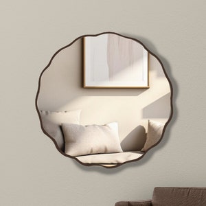 May include: A round, brown-framed mirror with a scalloped edge hangs on a light beige wall. The mirror reflects a room with a sofa and cushions.
