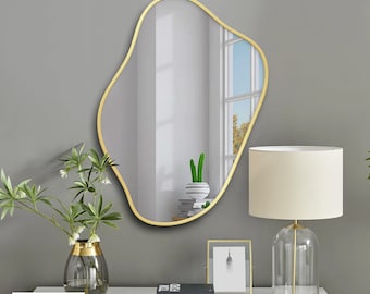 May include: An irregularly shaped mirror with a gold-colored frame hangs on a gray wall. A white table holds a lamp, a framed picture, and a vase with greenery. The mirror reflects a window and the text "BCD MIRROR".