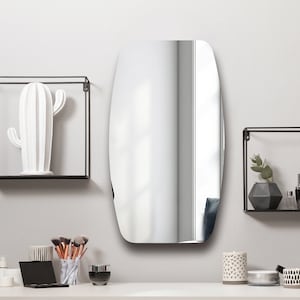 May include: A white ceramic cactus sculpture sits on a black metal shelf. A round, oval-shaped mirror hangs on the wall above a white countertop with makeup brushes, a black makeup palette, and a clear glass bottle. Two black metal shelves with decorative items are on the wall.