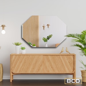 Puede incluir: Espejo octogonal con borde biselado, que refleja una pared de madera y una habitación con luces colgantes y plantas. El espejo está colgado sobre una cómoda de madera con un patrón de chevron.