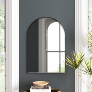 May include: An arched wall mirror with a dark gray frame hangs on a dark teal wall. The mirror reflects a bright window and a glimpse of the room. A small table below holds a wooden bowl and books.