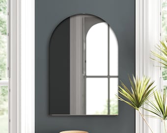 May include: An arched wall mirror with a dark gray frame hangs on a dark teal wall. The mirror reflects a bright window and a glimpse of the room. A small table below holds a wooden bowl and books.