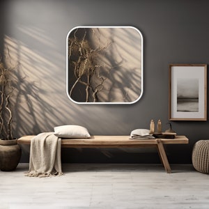 May include: A square mirror with rounded corners and a white frame hangs on a grey wall. The mirror reflects a dried branch against a light brown background. A wooden bench with a white throw blanket sits in front of the mirror.
