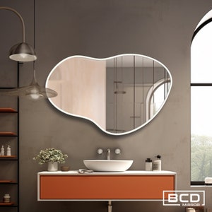 May include: A white, irregularly shaped bathroom mirror with a white frame, hanging on a gray wall above a white sink and orange countertop. The mirror reflects a bathroom with a window and a shelf.