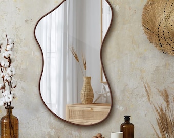Puede incluir: Un espejo de forma irregular con un marco marrón oscuro cuelga de una pared texturizada. El espejo refleja una habitación con un jarrón tejido con espigas de trigo, un mueble de madera y un sombrero de paja. El logotipo "BCD MIRROR" es visible.