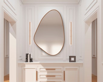 May include: An irregularly shaped mirror with a bronze frame hangs on a white wall. The mirror is above a white cabinet with bronze drawer pulls. Two vertical bronze lights flank the mirror. The room has white panelling and a light wood floor. The mirror is a decorative item.