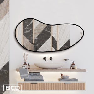 May include: A black framed, freeform shaped mirror with a geometric pattern of black, white, and beige marble tiles. The mirror is hanging above a white bathroom sink with a white countertop and wooden cabinets.
