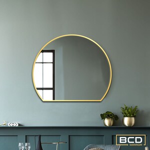 Peut inclure: Miroir rond avec un cadre doré, fixé sur un mur turquoise. Le miroir a une base plate et un sommet incurvé. Le texte "BCD MIRROR" est dans un rectangle doré en bas à droite.