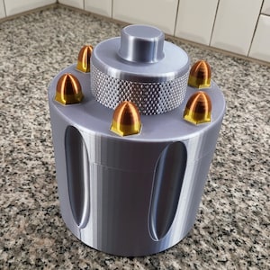 May include: A silver metal container with a textured surface and a round top. The container has six slots around the top, each with a gold-colored bullet-shaped object.