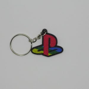 May include: A keychain featuring the PlayStation logo. The logo is in red, blue, green, and yellow, with a black outline. The keychain has a silver key ring and chain.