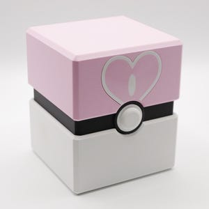 May include: A geometric box in pink, black, and white. The top pink section has a white heart design. A white circular button is centered on the front. The box is likely for storage or display.