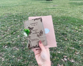 May include: A wooden wedding invitation featuring floral and butterfly motifs, the names "Paula and Patrick", and the date "May 21 2025". The invitation is tied with twine and decorated with dried flowers. A matching envelope is included.